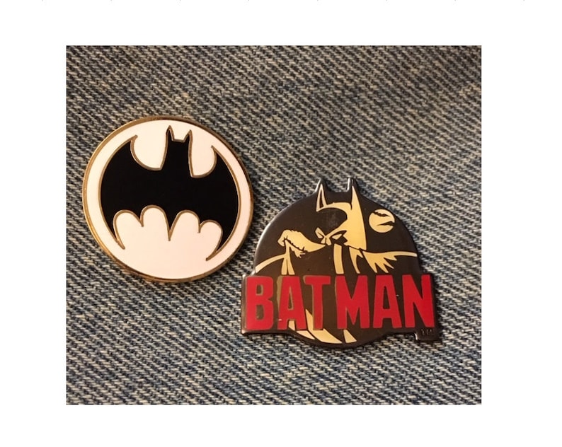 Op de afbeelding: Twee Batman-speldjes. E&eacute;n is een zwart-wit silhouet van het Batman-logo. De andere is een zwart-gouden silhouet van Batman met het woord "BATMAN" in rode letters.