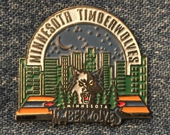 May include: A gold and green enamel pin featuring a wolf howling at the moon over a cityscape. The pin says "Minnesota Timberwolves" around the top and "Minnesota Timberwolves" at the bottom.