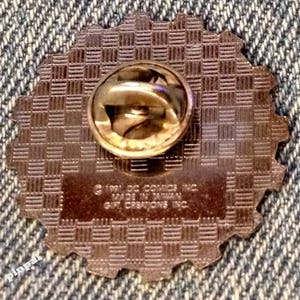 May include: The back of a gold-colored pin with a checkered pattern. The pin has a safety clasp and the text "(c) 1991 DC COMICS INC. MADE IN TAIWAN GIFT CREATIONS INC."