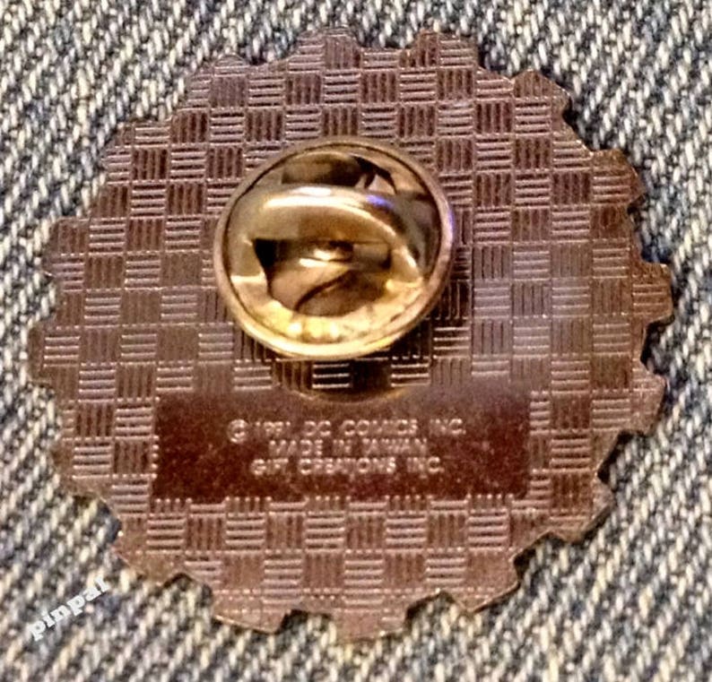 May include: The back of a gold-colored pin with a checkered pattern. The pin has a safety clasp and the text "(c) 1991 DC COMICS INC. MADE IN TAIWAN GIFT CREATIONS INC."