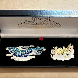 May include: A black presentation box containing two decorative pins. One pin shows a blue whale design with white clouds. The other pin depicts characters on a cloud. The box has gold lettering that reads "45 Years Of Magic".