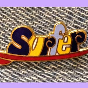 May include: A gold-coloured pin with the word "Surfer" in blue, yellow and white letters on a red surfboard. The pin is on a blue denim background.