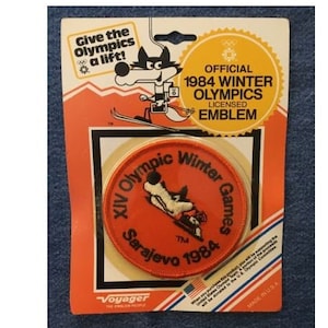 May include: A red and black embroidered patch with the text "XIV Olympic Winter Games Sarajevo 1984" and a cartoon wolf skiing. The patch is on a white background with a yellow border. The text "Official 1984 Winter Olympics Licensed Emblem" is on a yellow starburst.