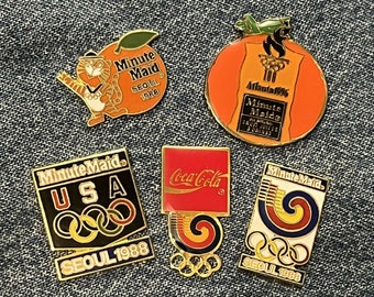 May include: A collection of six vintage enamel pins featuring the Minute Maid brand logo. The pins feature the Olympic rings and the years 1988 and 1996. The pins are in various colors, including orange, red, blue, yellow, and black.