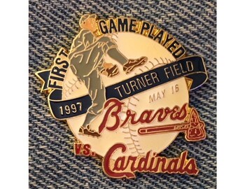May include: A gold-colored baseball pin commemorating the first game played at Turner Field on May 16, 1997. The pin features a baseball with a pitcher in action, the Atlanta Braves logo, and the text "First Game Played Turner Field May 16, 1997" and "Braves vs. Cardinals".