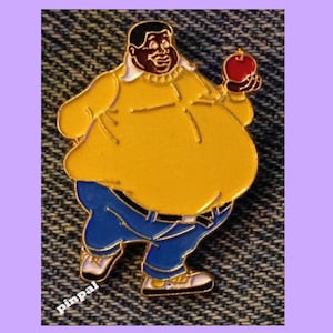 May include: A gold-coloured enamel pin depicting a cartoon character wearing a yellow jumper, blue jeans, and holding a red apple. The character has a large belly and a wide smile.