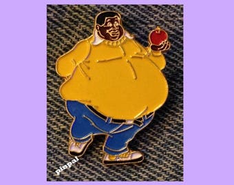 May include: A gold-colored enamel pin featuring a cartoon character wearing a yellow sweater, blue jeans, and holding a red apple. The character has a large belly and a wide smile.