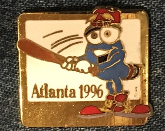 May include: Gold-colored pin featuring a cartoon character wearing a red and white hat, blue overalls, and red shoes. The character is holding a baseball bat and is depicted swinging at a baseball. The text "Atlanta 1996" is printed below the character.