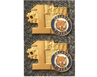 May include: Two gold-colored #1 Fan pins with a Detroit Tigers logo. The logo features a tiger head with orange and white stripes on a blue circle with the words "Detroit Tigers" in white.