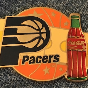May include: A gold-colored pin with a basketball, the word "Pacers", and a Coca-Cola bottle. The basketball is orange and black, and the bottle is red with a green cap.