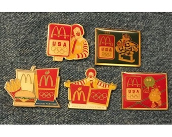 May include: Five gold pins featuring the McDonald's logo and the Olympic rings. The pins are red, yellow, and blue with the words "USA" and "Proud Partner" on them. One pin features a cartoon character wearing a red and yellow uniform.