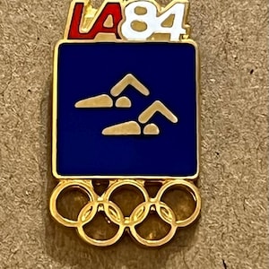 Könnte beinhalten: Goldfarbene Anstecknadel mit dem LA '84-Logo in Rot und Weiß. Die Nadel hat ein blaues Quadrat mit goldenen Schwimmer-Symbolen und den olympischen Ringen unten. Ein Vintage-Sammlerstück.