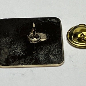 May include: A gold-colored metal pin with a black square back. The pin has a safety clasp on the back.