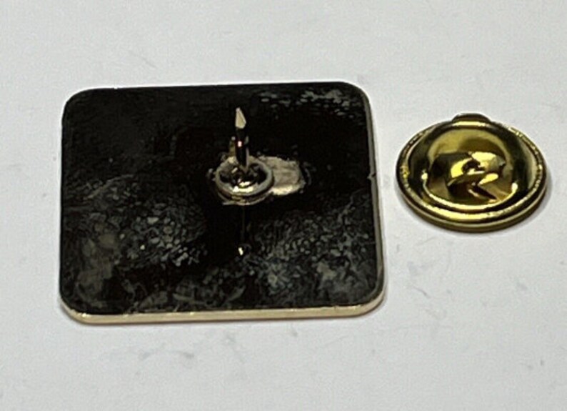 May include: A gold-colored metal pin with a black square back. The pin has a safety clasp on the back.