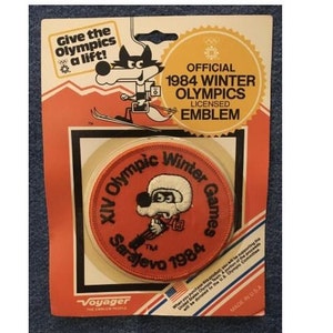 May include: A red and white embroidered patch featuring the text "XIV Olympic Winter Games Sarajevo 1984" and a cartoon wolf wearing a hat. The patch is packaged in a plastic bag with a card that reads "Official 1984 Winter Olympics Licensed Emblem".