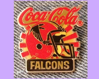 May include: A gold and red Coca-Cola pin featuring a black and yellow football helmet with a red and black bird on top. The pin says "FALCONS" in gold letters on a black background.