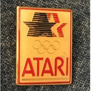 May include: A gold-colored pin with a red and black star design and the word "ATARI" in red letters. The pin also features five interlocking rings, a symbol of the Olympic Games.