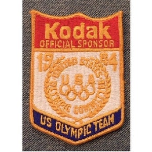 Puede incluir: Un parche bordado blanco y dorado con el texto "Kodak Patrocinador Oficial 1984 Equipo Olímpico de EE. UU.". El parche tiene un fondo rojo con la palabra "Kodak" en letras doradas. El parche presenta una corona dorada con cinco anillos entrelazados en el centro.