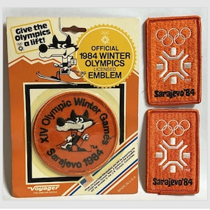 May include: A set of three orange and white embroidered patches featuring the 1984 Winter Olympics logo and the text "Sarajevo '84". The patches are on a white background with a red, white, and blue border.