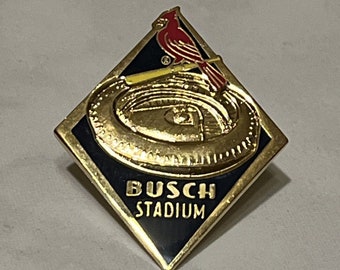 May include: A gold-colored pin with a black diamond-shaped background. The pin features a red cardinal perched on a baseball bat above a gold-colored baseball stadium with the text "Busch Stadium".