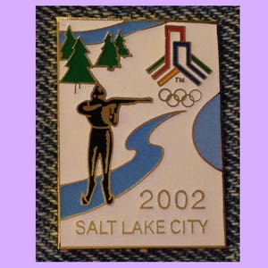 Può includere: Uno spillo in smalto bianco con bordo dorato. Lo spillo presenta una silhouette nera di un biatleta che spara con un fucile, una catena montuosa stilizzata blu e bianca e gli anelli olimpici. Il testo "2002 SALT LAKE CITY" è stampato in oro nella parte inferiore.