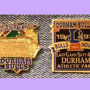 May include: Two gold-coloured baseball pins. The first pin features a detailed image of a baseball field with the text "Durham Athletic Park" and "Durham Bulls". The second pin features the text "Durham Bulls" and "1993 Bulls vs. 1993 Indians" with a bull mascot and the text "Last Game-Sept 4th Durham Athletic Park".