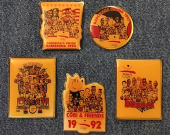 May include: Five gold-colored pins with images of basketball players. The pins feature the text "Olympic Champions", "America's Pride Barcelona 1992", "Barcelona Bound 1992", "Cobi & Friends 19-92", and "Dream Team".