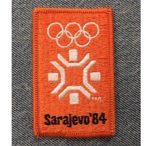 Könnte beinhalten: Orangener Stoff-Patch mit den Olympischen Ringen und einem Schneeflocken-Design. Der Text "Sarajevo '84" ist in Schwarz gestickt.