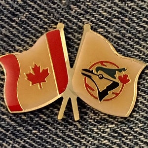 May include: A gold-toned pin depicting two crossed flags. One flag shows the Canadian maple leaf, the other a blue jay logo with a maple leaf. Both flags have a cream background with red and gold detailing.