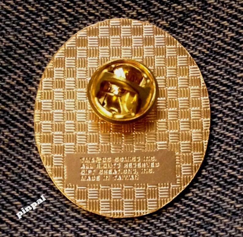 May include: The back of a gold-colored pin with a checkered pattern. The pin has a safety clasp and the text "TM & &copy; 2007,  Inc. All Rights Reserved. Gift Creations, Inc. Made in Taiwan."