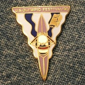 May include: A gold-coloured pin with a triangular shape. The pin features a white and blue design with a stylised image of a person in a canoe. The text "U.S. OLYMPIC FESTIVAL '90" is printed on the pin.