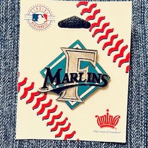 May include: A pin featuring the Miami Marlins logo, with a teal and silver design. The pin is on a cream-coloured card with red stitching and the MLB logo. The card also has the text "Genuine Merchandise".