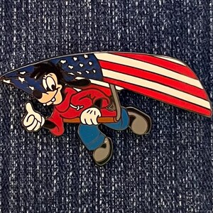 Puede incluir: Un pin colorido con un personaje de dibujos animados con una chaqueta roja y pantalones azules, sosteniendo una bandera estadounidense estilizada. El pin tiene un gesto de pulgar hacia arriba y está sobre un fondo de mezclilla azul oscuro.