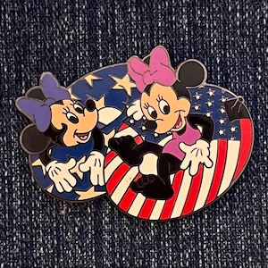 Puede incluir: Un pin colorido con dos ratones de dibujos animados, cada uno con un lazo y un vestido. El fondo está dividido en secciones de rayas rojas y blancas y azul con estrellas blancas. Los ratones están sonriendo.