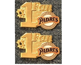 May include: Two gold-colored metal pins featuring the San Diego Padres logo. The pins are shaped like the number one with the word "Fan" above the logo. The logo is a white circle with the words "San Diego Padres Baseball Club" around the edge. The words "Padres" are in black letters with an orange outline.
