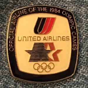 Könnte beinhalten: Eine goldene und schwarze Anstecknadel mit dem Text "Official Airline of the 1984 Olympic Games" um einen weißen Hintergrund mit dem United Airlines Logo, einem Stern und den olympischen Ringen.