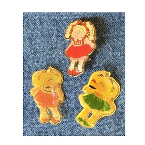 Puede incluir: Tres pines vintage de Cabbage Patch Kid. Los pines son de metal y tienen un fondo amarillo con una imagen de dibujos animados de un Cabbage Patch Kid. El primer pin presenta a una niña con un vestido rojo, el segundo pin presenta a una niña con un vestido rosa y el tercer pin presenta a una niña con un vestido verde.