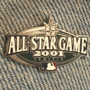 Peut inclure: Une épingle dorée avec un design vert et blanc. L'épingle présente le texte "All-Star Game 2001 Seattle" et la silhouette d'un joueur de baseball.
