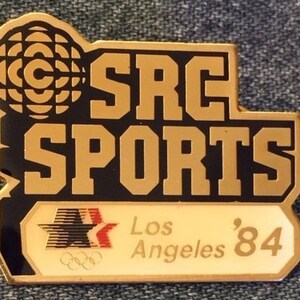 Könnte beinhalten: Eine goldene und schwarze Anstecknadel mit dem Text "SRC SPORTS" und "Los Angeles '84". Die Anstecknadel zeigt einen stilisierten Globus mit konzentrischen Kreisen und einen Stern mit rot-weiß-schwarzen Streifen.