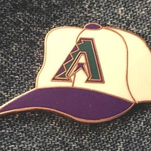 May include: A baseball cap pin with a white crown, purple bill, and a green and purple letter "A" in the center. The pin has a gold-colored outline and is set against a textured background.