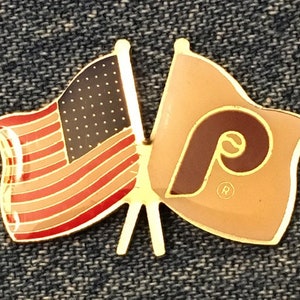 May include: Gold-colored pin with two flags crossed. The left flag is the American flag with red and white stripes and blue stars. The right flag is tan with a brown "P" in the center. The "P" has a registered trademark symbol.