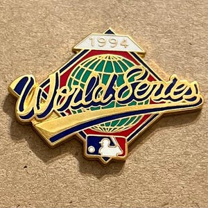 以下が含まれることがあります： 1994年のワールドシリーズの金色のピン。ピンには、スタイリッシュなフォントで「ワールドシリーズ」の文字と、中央に野球の地球儀のデザインが施されています。MLBのロゴが一番下にあります。