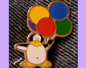 May include: A gold-colored penguin pin with a black beak and eyes, holding a bunch of colorful balloons. The balloons are red, yellow, blue, green, and purple.