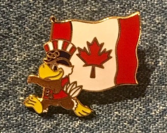 May include: A gold-colored pin featuring a cartoon eagle wearing a red, white, and blue hat and holding a Canadian flag. The eagle is running and the flag is waving.