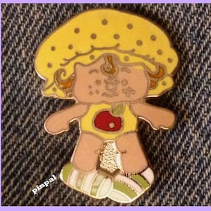 May include: A yellow pin with a cartoon image of a girl wearing a yellow hat with brown hair and a yellow dress with a red apple on the front. The girl is smiling and has brown eyes.