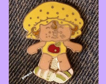 May include: A yellow pin with a cartoon image of a girl wearing a yellow hat with brown hair and a yellow dress with a red apple on the front. The girl is smiling and has brown eyes.