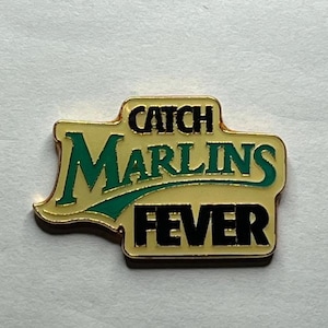 May include: A gold-coloured pin with a white background and the text "Catch Marlins Fever" in black and green lettering.