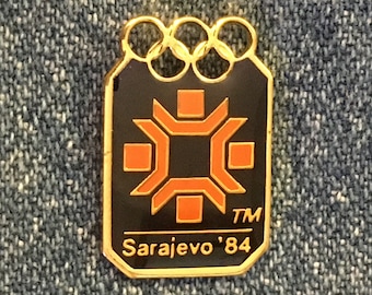May include: Gold-colored pin with the Olympic rings and a black and orange logo. The logo features a stylized geometric design. The text "Sarajevo '84" and "TM" are printed below the logo.