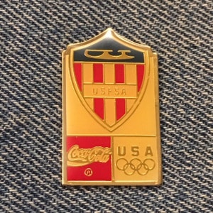 May include: Gold-colored pin with a shield design featuring red and white stripes, the text "USFSA", and a black and gold logo. The bottom of the pin features the Coca-Cola logo and the text "USA" with the Olympic rings.