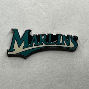 May include: A teal and white metal pin with the word "Marlins" in a stylised font.